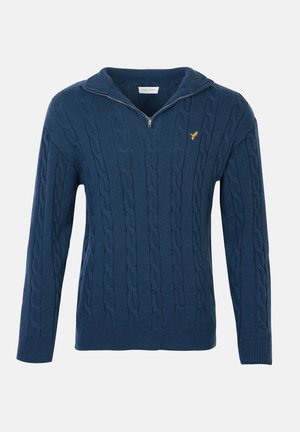 Suéter de punto azul con cuello de media cremallera y pequeño logo de un pájaro amarillo en el lado izquierdo del pecho, mangas largas y puños y dobladillo acanalados.