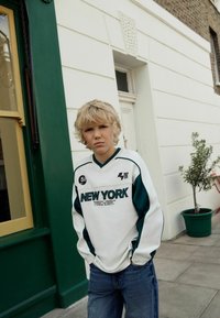 Biely a tyrkysový športový dres s nápisom "NEW YORK", tmavozelené akcenty, spárovaný s modrými džínami. Jednoduché pozadie s kvetináčom.