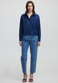 Navy suède blouse met lange mouwen, enkele borstzak; gecombineerd met hooggepaste blauwe jeans en bruine sandalen met straps.