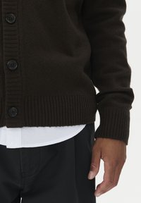 Brūns adīts kardigans ar ribotām piedurknēm un apakšmalu, kuram priekšā ir melnas pogas. Zem tā ir uzvilkta balta krekls.