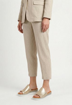 Person iført beige cropped bukser og matchende blazer med gyldne åbne slip-on platform sandaler, står på hvidt gulv.