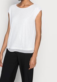 Top blanco de manga corta con un diseño texturizado y con lentejuelas, que presenta un escote redondo y un dobladillo en capas, combinado con pantalones negros.