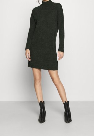 Kvinde iført en mørkegrøn ribstrikket minikjole med lange ærmer og sorte læderankelstøvler, stående mod en ensfarvet baggrund.