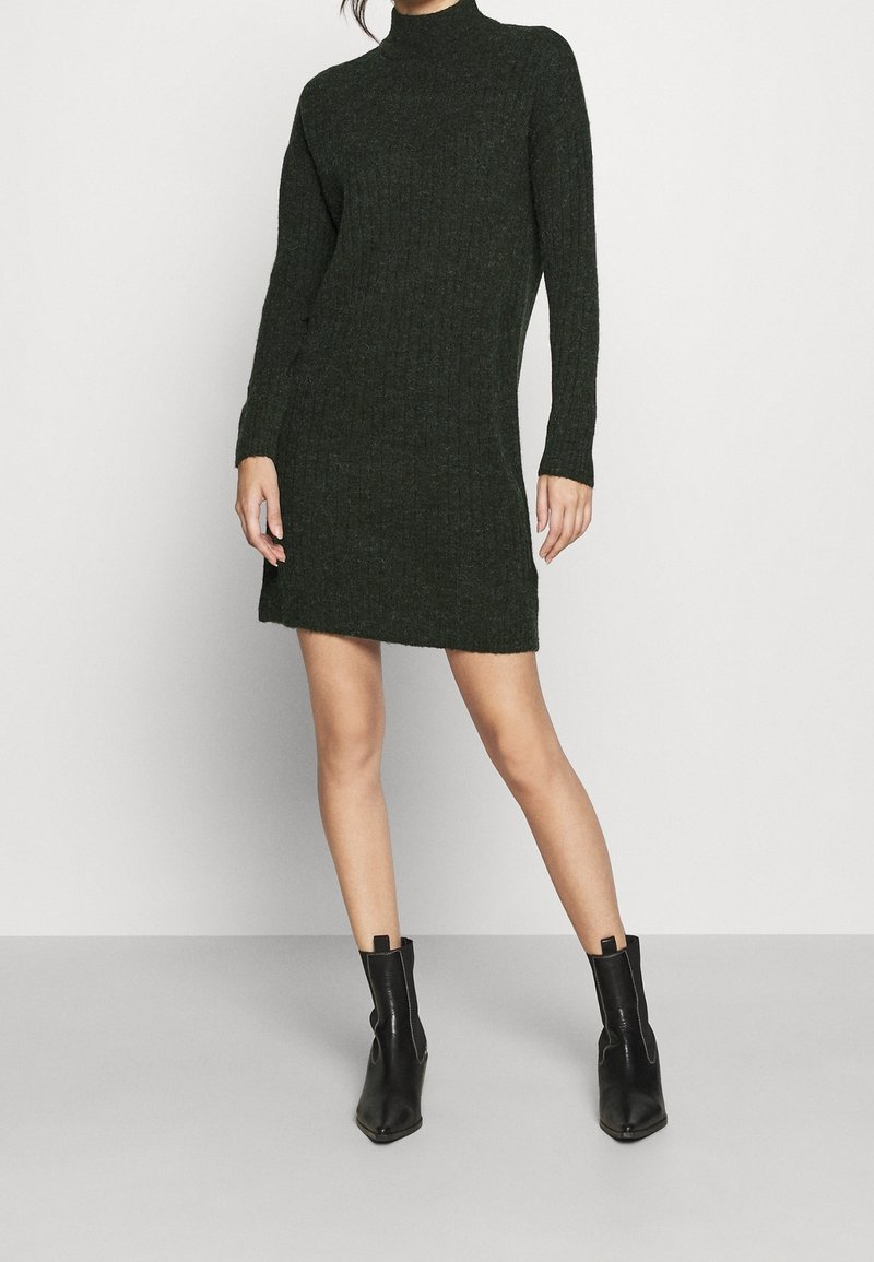 Femme portant une mini-robe en maille côtelée à manches longues de couleur vert foncé et des bottines en cuir noir, debout devant un fond uni.