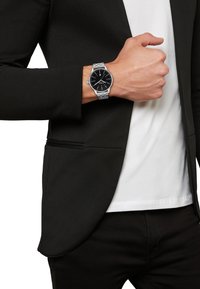 Montre en métal argenté avec un cadran noir, boîtier rond, index en chiffres romains, portée au poignet avec un blazer noir texturé et une chemise blanche en dessous.