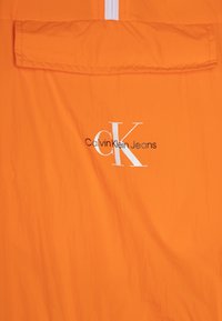 Kurtka Calvin Klein Jeans w jaskrawym pomarańczowym kolorze, charakteryzująca się gładką teksturą, z przodu zapinaną na zamek oraz z nadrukowanym logo w białym i czarnym kolorze.