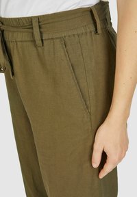 Olivgrüne Leinenhose mit gebundenem Bündchen, seitlichen Taschen und einer leichten Textur. Sichtbare Nähte entlang der Kanten.