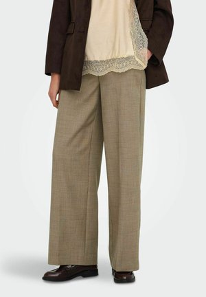 Personne portant un pantalon beige à jambes larges, un haut crème orné de dentelle, un blazer marron foncé et des chaussures en cuir marron foncé, debout les mains détendues.