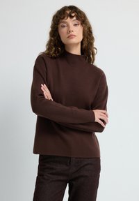 Vrouw met krullend haar die een donkerbruine lange mouwen trui en bijpassende broek draagt, staand met gekruiste armen tegen een effen achtergrond.