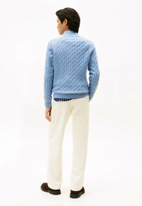 Mann steht mit dem Rücken zur Kamera, trägt einen hellblauen Zopfmuster-Pullover, cremefarbene Hose mit Taschen und dunkelbraune Schuhe vor einem weißen Hintergrund.