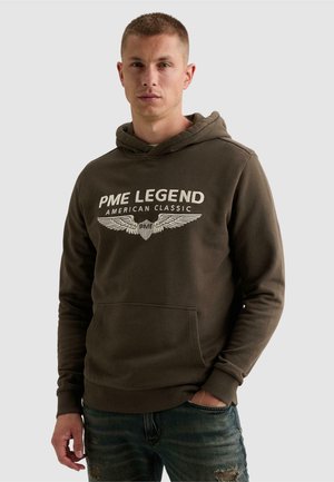 Młody mężczyzna w ciemnozielonej bluzie z kapturem z "PME Legend American Classic" i emblematem ze skrzydłami, w zestawieniu z postrzępionymi niebieskimi dżinsami.
