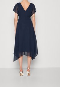 Mörkblå klänning med V-rygg, korta volangärmar och asymmetrisk fåll. Tillverkad av lättviktsmaterial med en flödande silhuett.