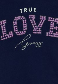 Felpa blu navy con "TRUE LOVE" in gemme viola, contornata da cuciture argentate, e "Guess" in un font corsivo sottostante. Dettagli testurizzati.