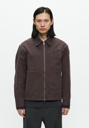 Brown cotton jacket with a collar, front zipper, and two large pockets. Features minimal seams and a textured finish.