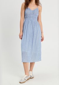 Robe midi rayée bleue avec fines bretelles et taille élastique, en tissu léger. Associée à des chaussures de sport argentées.