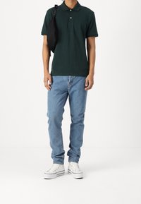 Mladý člověk nosící tmavě zelené polo triko, světle modré džíny, bílé tenisky a černou krosbody tašku přehozenou přes jedno rameno.