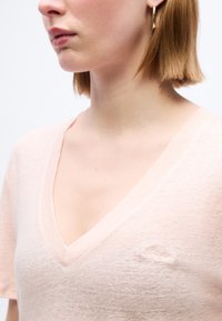 Γυναίκα με κοντά ξανθά μαλλιά που φοράει χρυσό σκουλαρίκι κρίκο και ανοιχτόροζ μπλούζα με V λαιμόκοψη και διακριτικό κεντημένο λογότυπο στο στήθος.