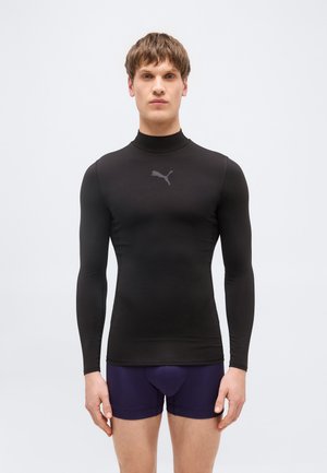 Férfi fekete, hosszú ujjú, testhez simuló Puma logós pólóban és sötétlila boxeralsóban, egyszínű háttér előtt áll.