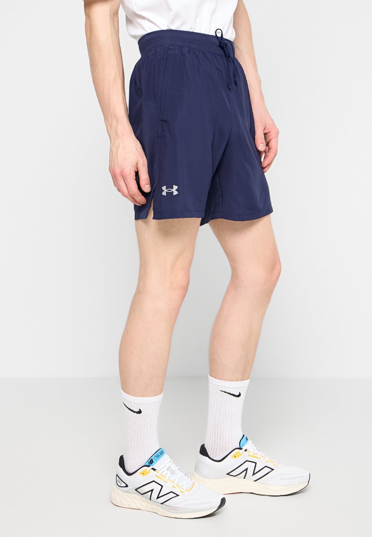 Hombre vistiendo pantalones cortos atléticos azul marino de Under Armour, calcetas blancas de Nike y zapatillas para correr blancas de New Balance frente a un fondo blanco liso.