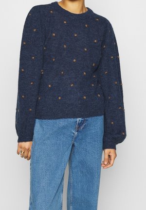 Vrouw draagt een marineblauwe trui met kleine bruine stippen en opbollende lange mouwen, gecombineerd met lichtblauwe jeans met hoge taille.