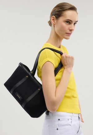 Mujer con camiseta amarilla de manga corta y pantalones blancos lleva un bolso tote negro de Trussardi sobre el hombro contra un fondo liso.