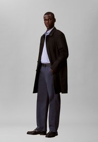 Άνδρας που στέκεται με τα χέρια στις τσέπες, φορώντας μαύρο παλτό, ανοιχτό μπλε πουκάμισο, γκρι παντελόνι, μαύρη ζώνη και μαύρα παπούτσια, σε γκρι φόντο.