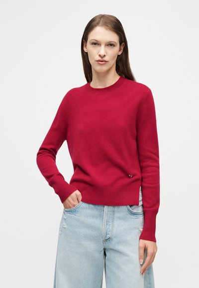 Γυναίκα με μακριά καστανά μαλλιά, ντυμένη με κόκκινο πουλόβερ με στρογγυλή λαιμόκοψη και ανοιχτό μπλε ψηλόμεσο τζιν, στέκεται με το ένα χέρι στην τσέπη μπροστά σε λευκό φόντο.
