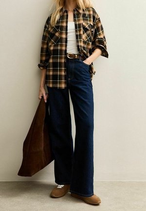 Vrouw die een bruin geruit overhemd draagt over een wit topje, donkere wijde spijkerbroek, bruine riem en schoenen, en een grote bruine tas vasthoudt.