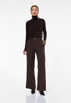 Kobieta stojąca z jedną ręką w kieszeni spodni, ubrana w czarny golf, brązowe szerokie spodnie, czarny pasek i czarne szpiczaste szpilki.