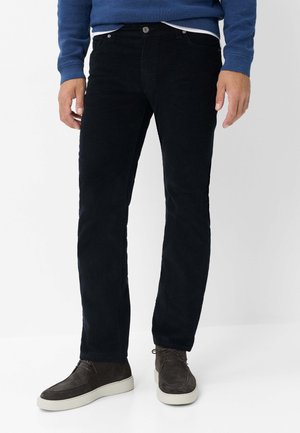 Uomo con pantaloni neri, scarpe stringate in camoscio grigio scuro con suole bianche e maglione blu a maniche lunghe, in piedi davanti a uno sfondo bianco.