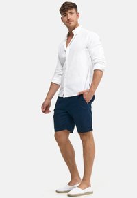 Weißes Leinenhemd, marineblaue Shorts und weiße Espadrilles. Das Hemd hat einen Button-Down-Kragen und lange Ärmel. Überall glatte Texturen.