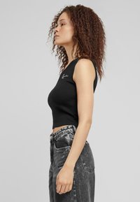 Svart ribbestrikket crop-top med ermeløst design, med et minimalt brodert logo. Kombinert med høy midje, falmede svarte jeans.