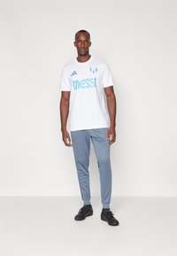 Hvid t-shirt med lys blå "MESSI" grafik og tre blå striber på skuldrene, parret med lysegrå joggers og sorte sneakers.