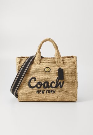 Gevlochten beige Coach New York tote tas met zwart logo, dubbele handvatten, vooraan draaivergrendelingssluiting en een zwarte gestreepte schouderriem.