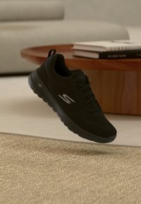 Baskets Skechers noires flottant au-dessus d'un tapis beige, avec une table basse en bois et des livres empilés flous en arrière-plan.