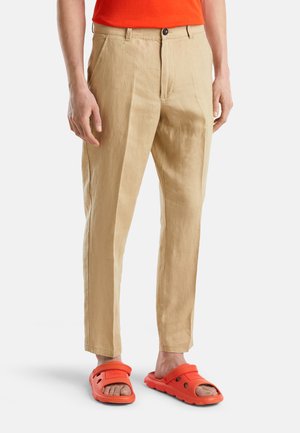 Mężczyzna noszący beżowe lniane spodnie, pomarańczowe sandały i pomarańczową koszulę, widoczny od pasa w dół na białym tle.