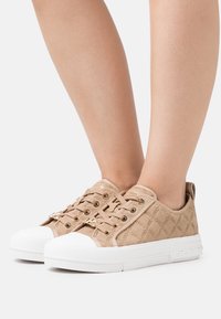 Baskets en toile beige avec un motif texturé, des oeillets dorés et des semelles en caoutchouc blanches. Présentent un accent logo sur le côté.