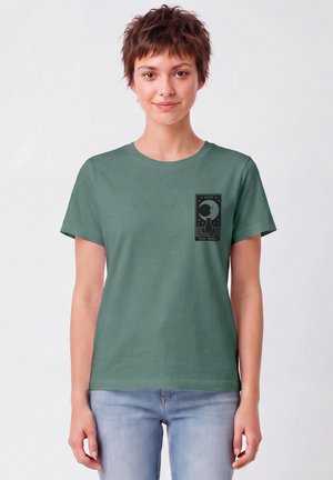 Jeune femme aux cheveux courts portant un t-shirt vert avec un motif chat noir et lune étiqueté "The Moon" sur la poitrine, associé à un jean clair.