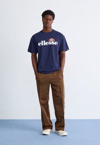Jūras zila kokvilnas t-kreklis ar baltu "ellesse" logo un sarkanas/baltas akcentiem, apvienots ar brūnām biksēm. Modelis stāv uz zila grīdas fona.