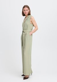 Jumpsuit senza maniche di colore verde chiaro con colletto, vita cintata, tasche laterali e vestibilità comoda; abbinato a scarpe a punta di colore marrone scuro.
