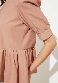 Top peplum rosa pallido con maniche corte a palloncino, tessuto in cotone morbido, vita arricciata e superficie liscia. Nessun motivo o accento particolare.