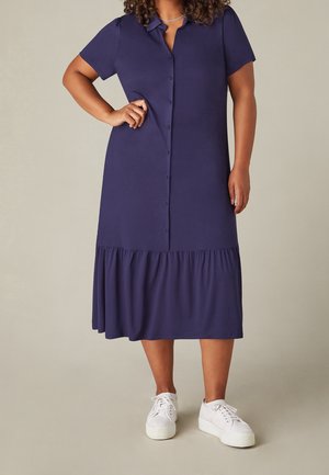 Femme portant une robe midi bleu marine à boutons, manches courtes, ourlet volanté et baskets blanches, debout avec une main sur la hanche.