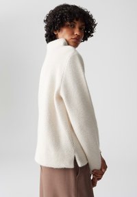 Maglione bianco texturizzato con colletto alto e spacchi laterali, indossato sopra un tessuto marrone. Maniche a tre quarti e vestibilità comoda.