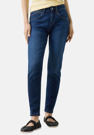 Persoon draagt donkerblauwe slim-fit jeans, een geel gestreept shirt en zwarte platte schoenen met ruches tegen een witte achtergrond.
