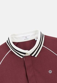 Polo bordeaux avec un col blanc à rayures noires, design à boutons et un petit logo sur la poitrine. Texture en coton lisse.