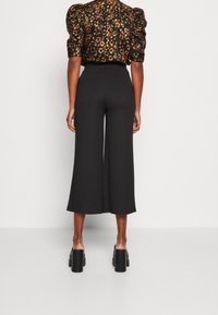 Pantalon noir à jambes larges et taille haute, fabriqué dans un tissu lisse. Le modèle porte des chaussures noires à talons carrés, présentant un design minimaliste.