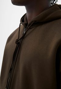 Felpa con cappuccio marrone caratterizzata da una texture liscia, con cordini regolabili e chiusura con zip, posizionata all'altezza della spalla e del collo.