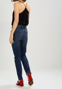 Mujer que lleva vaqueros oscuros, una blusa sin mangas negra con tirantes finos y sandalias de tacón cuadrado rojas, de pie frente a un fondo liso.