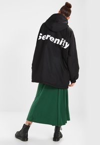 Chaqueta negra de gran tamaño con el texto blanco "Serenity" en la parte posterior, combinada con una falda plisada verde y botas negras con cordones.
