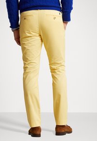 Person som bär gula slim-fit-byxor, blå tröja och bruna mockaskor, står med händerna i fickorna, vänd med ryggen mot kameran.